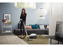 Charger l&#39;image dans la galerie, Une femme passe l&#39;aspirateur dans un salon avec un canapé gris, un tapis vert et des objets décoratifs. Une grande lampe blanche pend.
