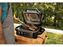 Charger l&#39;image dans la galerie, Une personne grillant des steaks sur un barbecue extérieur. Il y a une table en bois et un fond vert.
