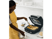Charger l&#39;image dans la galerie, Une femme avec un tablier jaune utilise des pinces pour déplacer de la nourriture sur un grill.
