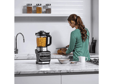 Charger l&#39;image dans la galerie, Une femme en chemise verte prépare de la nourriture avec un mixeur sur un comptoir.
