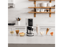 Charger l&#39;image dans la galerie, Un appareil Ninja avec de la glace et des milkshakes sur un comptoir blanc, dans une cuisine. Étagères et ustensiles en arrière-plan.
