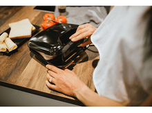 Charger l&#39;image dans la galerie, Une femme utilise un appareil à sandwich noir sur un comptoir en bois. Pain, tomates et fromage sont à proximité.
