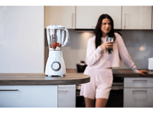 Charger l&#39;image dans la galerie, Mixeur blanc avec fraises rouges et banane sur comptoir. Femme en arrière-plan.
