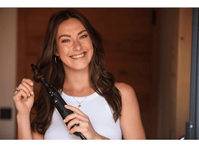 Charger l&#39;image dans la galerie, Femme souriante tenant un outil de coiffure noir, portant un haut blanc. Porte en bois en arrière-plan.
