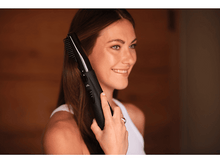 Charger l&#39;image dans la galerie, Femme se brossant les cheveux avec un lisseur noir, souriante. Fond en bois brun.
