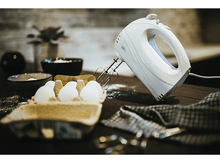 Charger l&#39;image dans la galerie, Un batteur à main blanc avec des œufs dans une boîte et de la farine sur une surface en bois.
