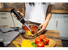 Charger l&#39;image dans la galerie, Une personne utilisant un mixeur plongeant noir dans une casserole de sauce tomate. Il y a des tomates, des pâtes et du basilic à proximité.
