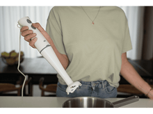 Charger l&#39;image dans la galerie, Une personne tient un mixeur plongeant blanc au-dessus d&#39;une casserole en inox sur une table.
