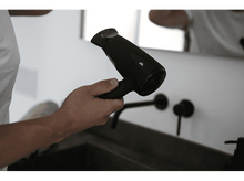 Charger l&#39;image dans la galerie, Une main tenant un sèche-cheveux noir près d&#39;un lavabo et d&#39;un miroir.
