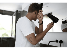 Charger l&#39;image dans la galerie, Homme se coiffant avec un peigne et un sèche-cheveux, face à un miroir, dans une salle de bain.
