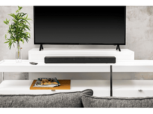 Charger l&#39;image dans la galerie, Une barre de son noire est située sous un téléviseur sur une table blanche. Une plante et un magazine sont à proximité.
