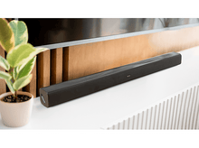 Charger l&#39;image dans la galerie, Barre de son noire sur une surface blanche, avec une plante et une télévision en arrière-plan.
