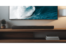 Charger l&#39;image dans la galerie, Barre de son noire sur une étagère en bois sous une TV montrant des vagues. Livres et lampe visibles.
