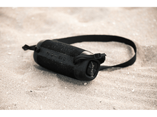 Charger l&#39;image dans la galerie, Un haut-parleur portable noir avec une sangle sur une plage de sable. Le haut-parleur est cylindrique.
