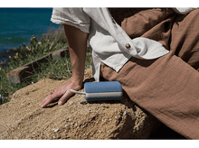 Charger l&#39;image dans la galerie, Femme avec haut-parleur bleu. Fond de plage. Vêtements beiges et blancs.
