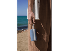 Charger l&#39;image dans la galerie, Main tenant un haut-parleur bleu. Fond de plage. Pantalon beige et chemise blanche.
