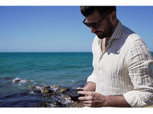Charger l&#39;image dans la galerie, Homme barbu portant des lunettes de soleil tenant un haut-parleur noir près de la mer.
