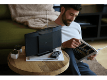 Charger l&#39;image dans la galerie, Un homme lisant un magazine à côté d&#39;un haut-parleur noir et de petites plantes.
