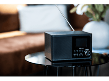 Charger l&#39;image dans la galerie, Une radio noire sur une table, avec un vase blanc et un canapé marron en arrière-plan.
