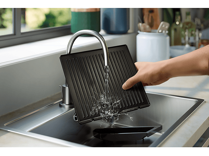 Une poêle grill noire est lavée sous un robinet. L'eau éclabousse.