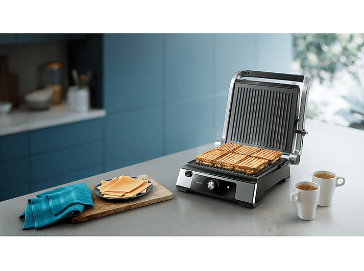 Sandwiches sur un grill, planche à découper en bois avec du fromage et deux tasses de thé. Une serviette bleue est à proximité.