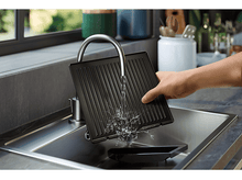 Charger l&#39;image dans la galerie, Une personne lave une plaque de cuisson noire dans un évier en acier inoxydable avec de l&#39;eau courante.
