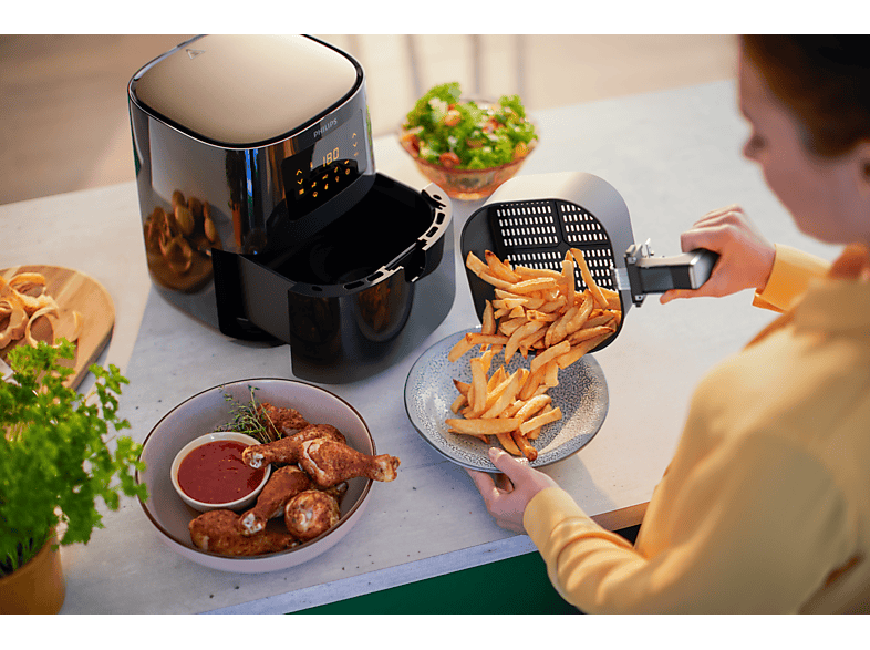 Friteuse à air avec des frites versées sur une assiette. Poulet et salade à proximité. Une personne tient.