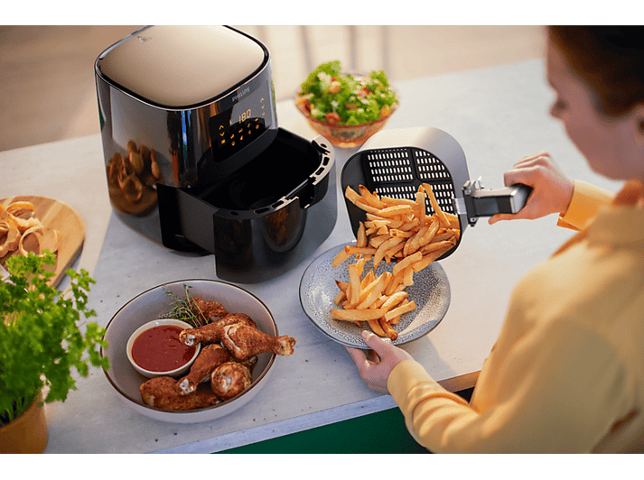 Friteuse à air avec des frites versées sur une assiette. Poulet et salade à proximité. Une personne tient.