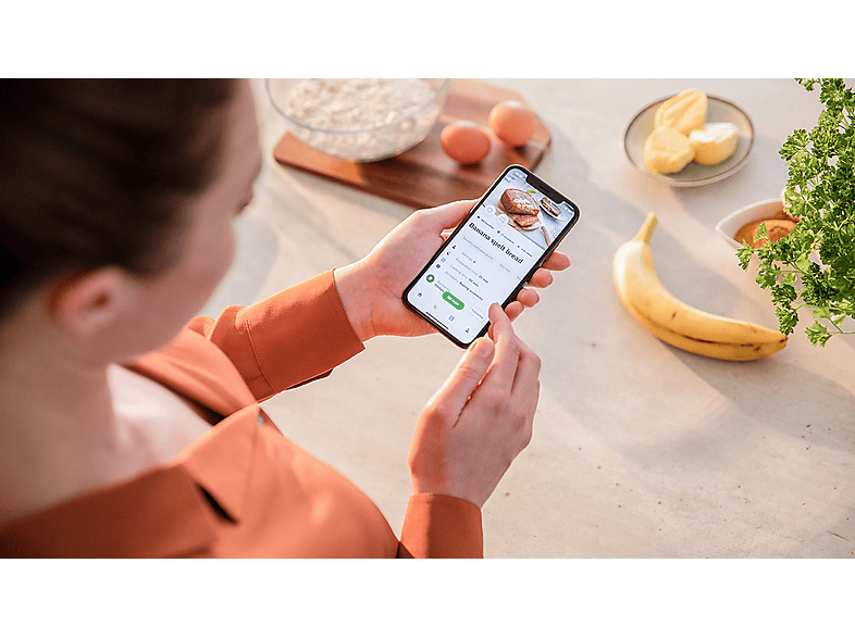 Une femme tenant un téléphone avec une recette, avec de la nourriture autour.