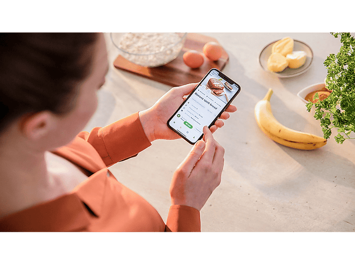 Une femme tenant un téléphone avec une recette, avec de la nourriture autour.