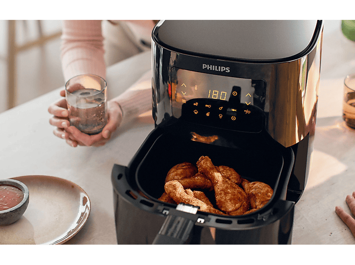 Friteuse à air avec des pilons de poulet frits dans le panier, personne tenant un verre.
