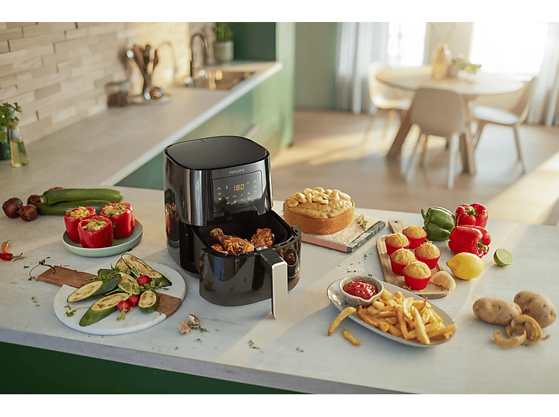 Friteuse à air noire avec aliments, entourée d'aliments cuits et frais sur un comptoir de cuisine.