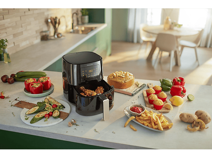 Friteuse à air noire avec aliments, entourée d'aliments cuits et frais sur un comptoir de cuisine.