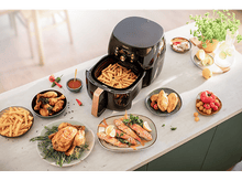 Charger l&#39;image dans la galerie, Friteuse à air avec des aliments cuits sur des assiettes, des légumes, du citron et un verre d&#39;eau, sur une table.
