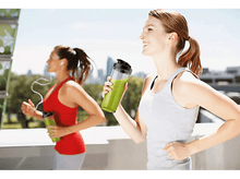 Charger l&#39;image dans la galerie, Deux femmes courent dehors, une avec un smoothie vert. L&#39;autre a des écouteurs.
