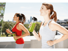 Charger l&#39;image dans la galerie, Deux femmes courant avec des smoothies verts dans des contenants, journée ensoleillée, scène extérieure, mode de vie sain.
