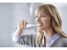 Charger l&#39;image dans la galerie, Femme se brossant les dents avec une brosse à dents électrique blanche. Elle sourit, a les cheveux blonds.
