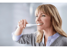 Charger l&#39;image dans la galerie, Femme se brossant les dents avec une brosse à dents électrique blanche. Souriante et regardant sur le côté.

