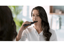 Charger l&#39;image dans la galerie, Femme se brossant les dents avec une brosse à dents électrique bleu foncé devant un miroir.
