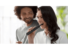 Charger l&#39;image dans la galerie, Femme se brossant les dents, souriante. Homme en arrière-plan souriant. Salle de bain blanche. Brosse noire.
