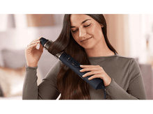 Charger l&#39;image dans la galerie, Femme utilisant un styler capillaire Philips noir sur des cheveux bruns. Arrière-plan flou.
