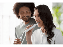 Charger l&#39;image dans la galerie, Une femme se brosse les dents avec une brosse à dents électrique, un homme sourit en arrière-plan.

