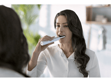 Charger l&#39;image dans la galerie, Une femme se brosse les dents en se regardant dans un miroir. Une brosse à dents électrique blanche est utilisée.

