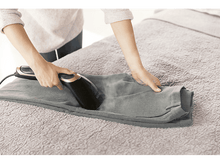 Charger l&#39;image dans la galerie, Une personne repasse un pantalon gris sur un lit avec un défroisseur à main. La scène est en intérieur.
