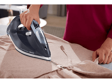 Charger l&#39;image dans la galerie, Femme repassant une veste. Un fer à repasser bleu foncé est utilisé sur un vêtement marron clair. Surface en bois.
