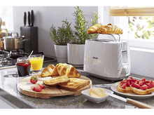 Charger l&#39;image dans la galerie, Un grille-pain blanc avec des croissants dessus et une table avec le petit-déjeuner.
