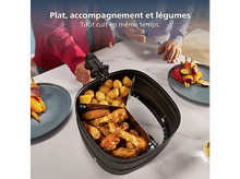 Charger l&#39;image dans la galerie, Une friteuse à air avec des pommes de terre, des ailes de poulet et des légumes. Deux personnes se tiennent au-dessus de la friteuse. Des assiettes avec de la nourriture de chaque côté.
