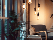 Charger l&#39;image dans la galerie, Scène d&#39;intérieur avec des ampoules suspendues, une sculpture de tête en bois et un fauteuil sombre.
