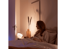 Charger l&#39;image dans la galerie, Une femme au lit regarde une lampe sur une table de chevet, avec une peinture en arrière-plan.
