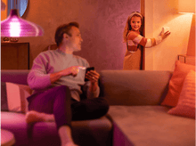 Charger l&#39;image dans la galerie, Un homme sur un canapé utilise un téléphone, une fille près d&#39;une porte allume une lumière.
